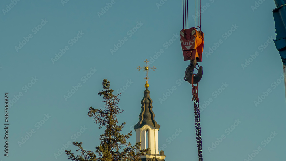Red hook of a crane on the background of the tower church with orthodox cross. Construction site.