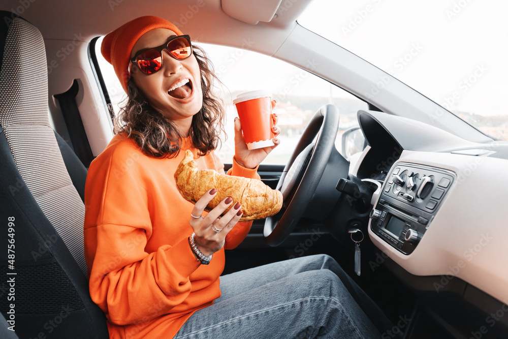 A happy young girl driver is drinking coffee and eating a fresh croissant while driving a car. The concept of ordering fast food delivery and dangerous driving