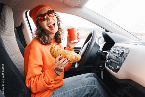 A happy young girl driver is drinking coffee and eating a fresh croissant while driving a car. The concept of ordering fast food delivery and dangerous driving