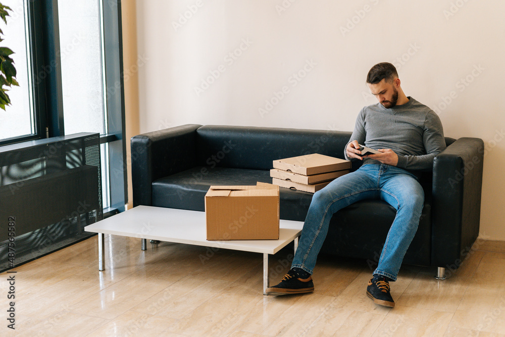 Delivery man holding using payment POS terminal waiting customer, sitting on sofa with boxes pizza and cardboard box parcel in hall of apartment or office building. Concept of online shopping.