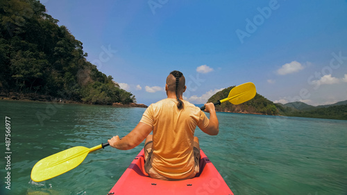 Strong man with thin plait hairstyle rows pink plastic kayak putting up paddle on sea against hills with wild jungles and blue sky. Young guy is sailing on kayak in ocean. Traveling to tropics. © ivandanru