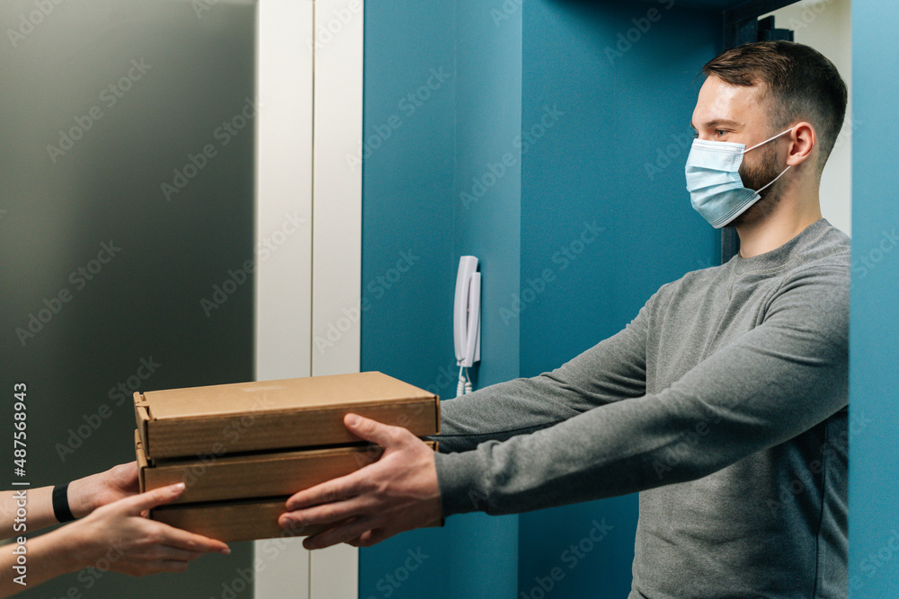 Side view of courier male wearing medical face mask delivering boxes with hot pizza to unrecognizable female customer on doorstep at home. Woman client receiving boxes with food from deliveryman. Side view of courier male wearing medical face mask delivering boxes with hot pizza to unrecognizable female customer on doorstep at home. Woman client receiving boxes with food from deliveryman.