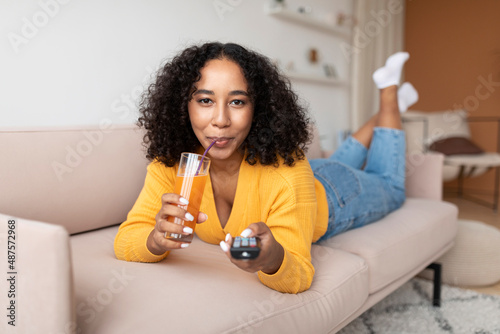 Pretty young black woman lying on sofa with TV remote and drinking juice, switching channels at home