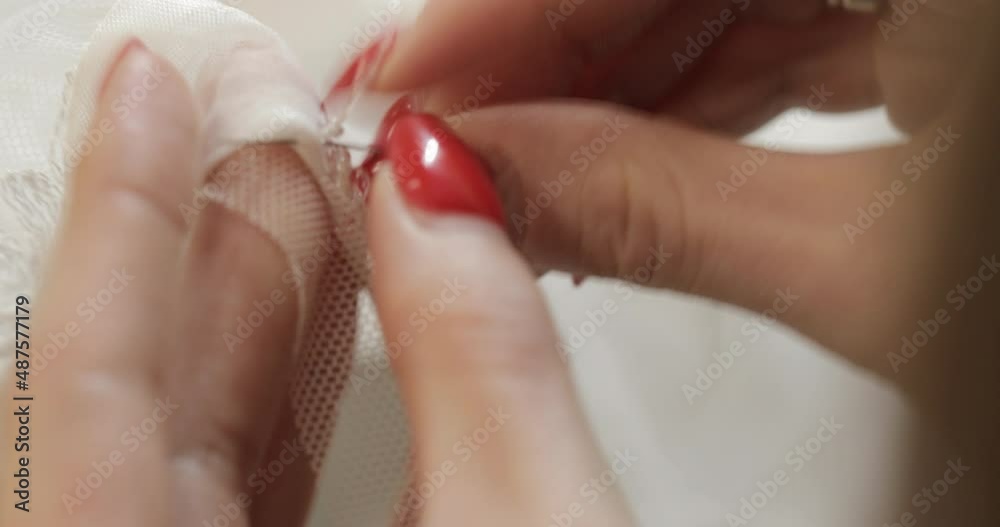 a seamstress in black clothes with a red manicure and a ring on her finger embroider with beads a detail of a wedding dress with a needle on a white transparent fabric with patterns. macro view. bokeh a seamstress in black clothes with a red manicure and a ring on her finger embroider with beads a detail of a wedding dress with a needle on a white transparent fabric with patterns. macro view. bokeh