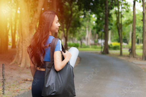 Beautiful woman wearing blue workout clothes holding rolled-up exercise mat in the park evening time. She is happy to go to the exercise.