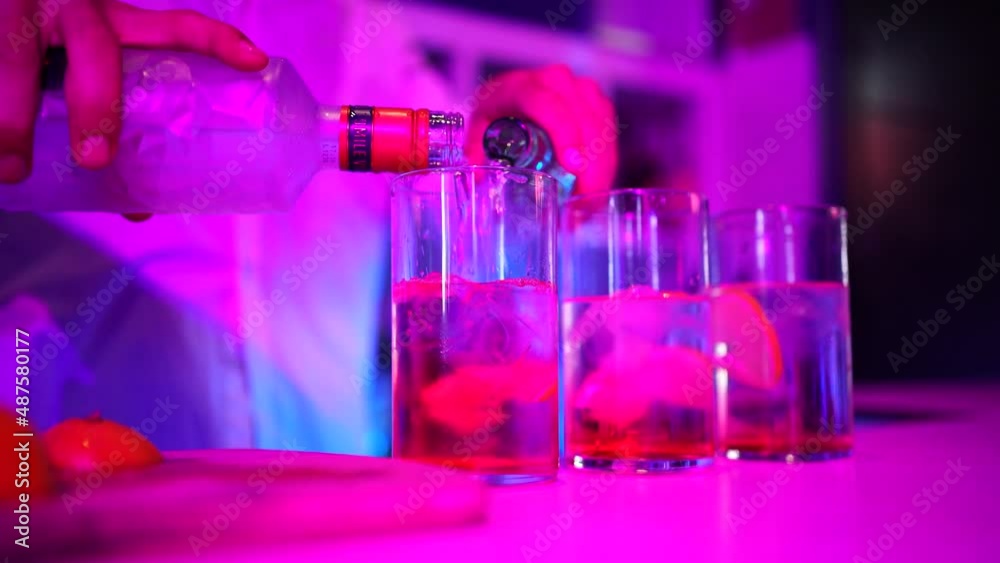 Se sirve una copa de ginebra con un refresco en la barra de una discoteca con luces de neón. El camarero llevo camisa blanca, se aprecian las manos con las mangas. Se ven 3 vasos con hielos y limón. 