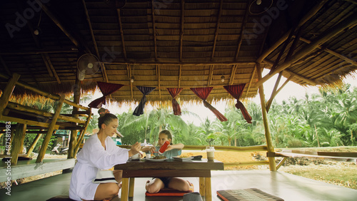 The happy family eat and drink tasty beverages spending time in local floating cafe on water. Mother and daughter having breakfast outdoors. Woman and a child in a cafe. Tropical view © ivandanru