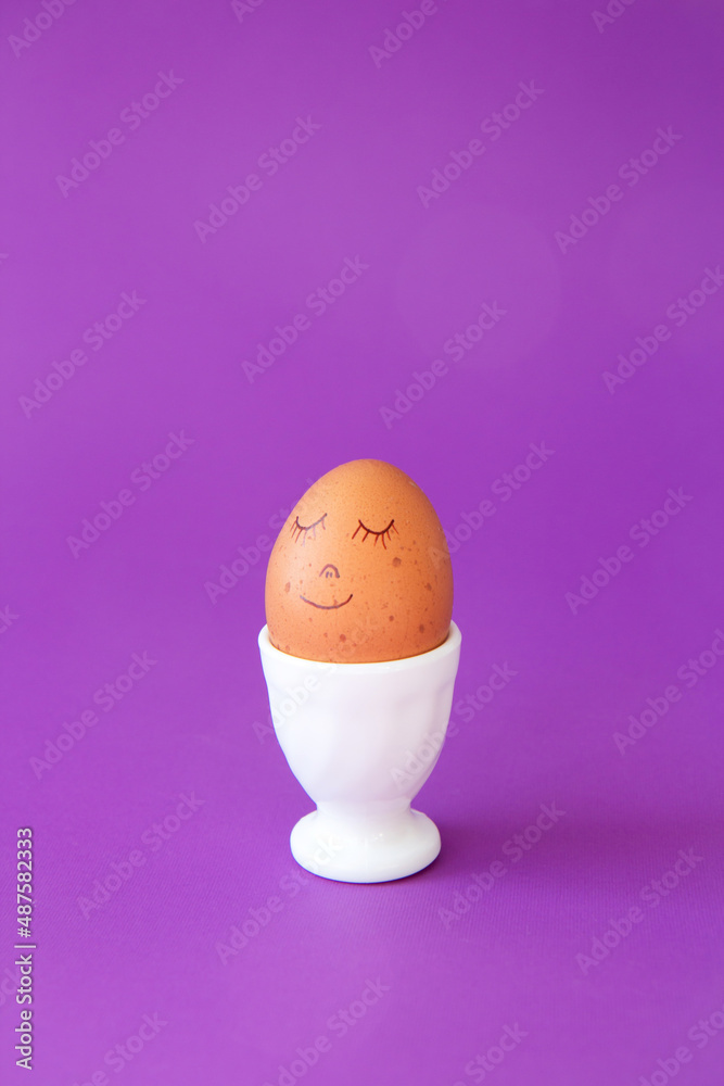 Easter smiling egg in egg cup. Easter holiday concept with cute handmade egg. Cute face egg on a violet periwinkle Easter smiling egg in egg cup. Easter holiday concept with cute handmade egg. Cute face egg on a violet periwinkle