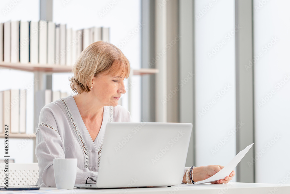 Portrait of woman checking their bills, Mature casual woman reading documents and working on a laptop in living room Portrait of woman checking their bills, Mature casual woman reading documents and working on a laptop in living room
