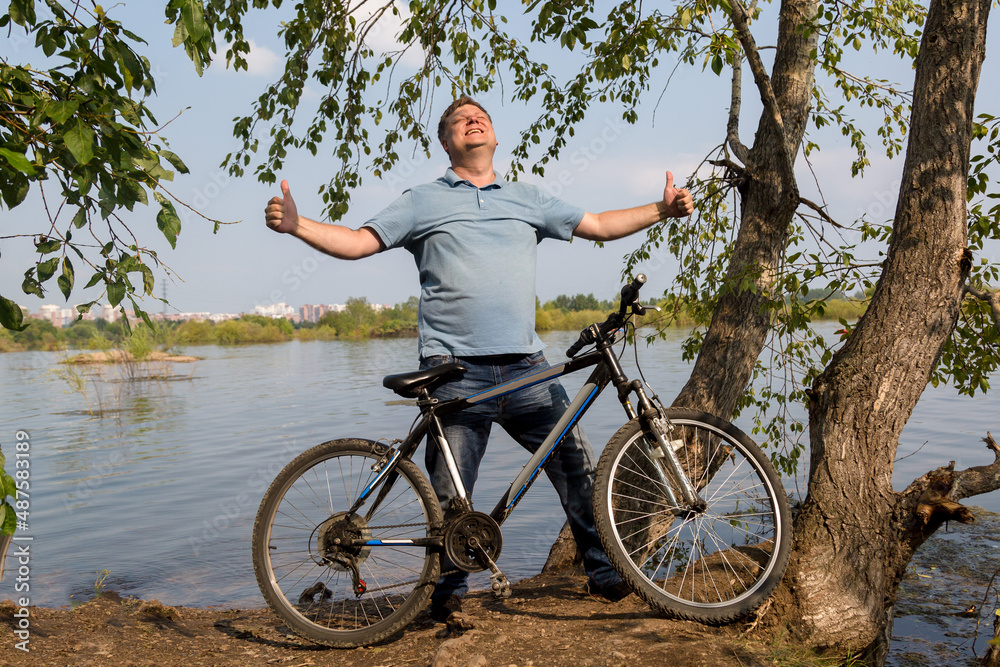 A tall man with a bicycle river rejoices and enjoys a sunny day with his hands raised to the sky.