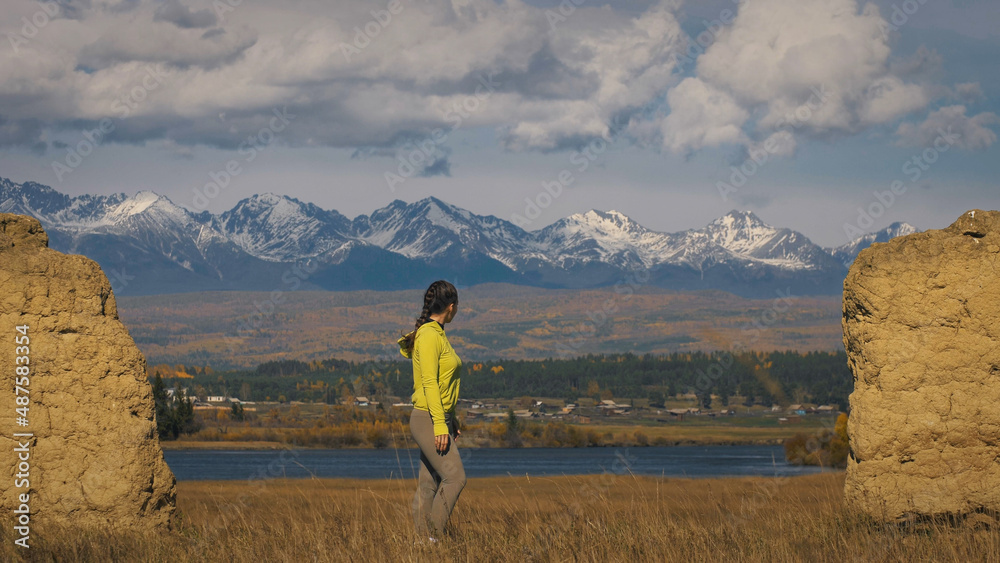 The woman in yellow green sportswear. The traveler near old stone enjoying highland landscape. Tourist are walking against the backdrop of snow-capped mountains. Strong wind.