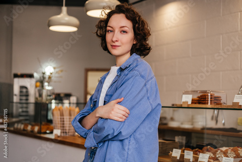 Smiling young woman bakery shop owner standing in front of showcase with pastry products. Small business concept