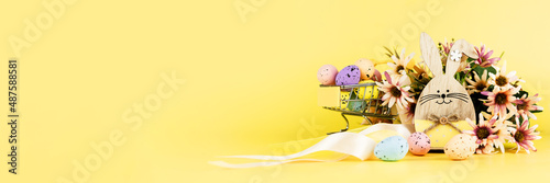 Photography Colorful easter quail eggs in shopping cart, wooden bunny and spring chrysanthemum flowers on yellow background
