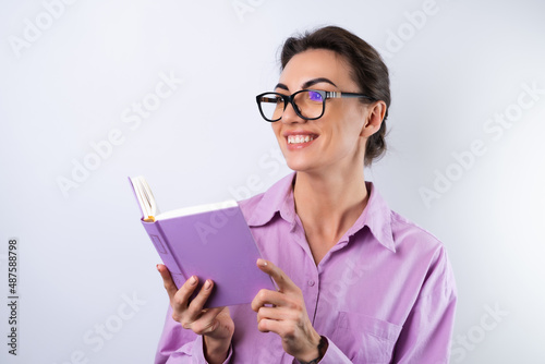 A young woman in a lilac shirt on a white background holds a book in her hands in glasses for vision