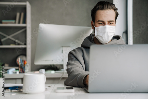 Young man in protective mask working with laptop at office
