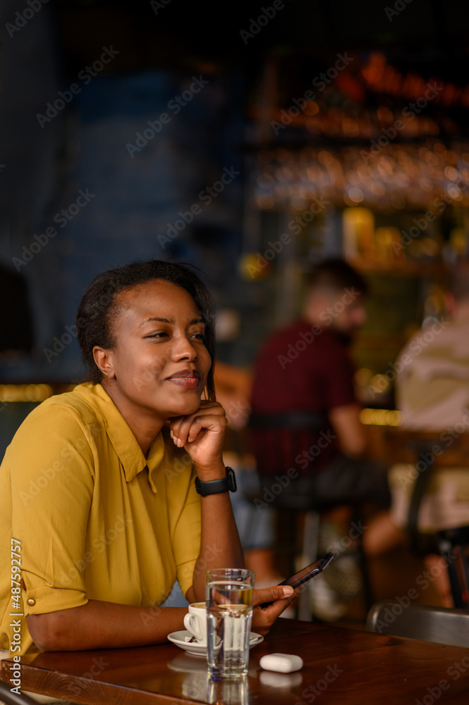African american woman using a smartphone and drinking coffee while sitting in a cafe