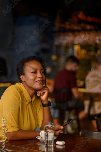 African american woman using a smartphone and drinking coffee while sitting in a cafe
