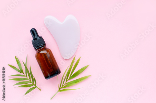 Cosmetic glass bottle with pipette, moisturizing gel guasha on pink background with tropical leaf. Skincare beauty product. Flat lay, top view, copy space.
