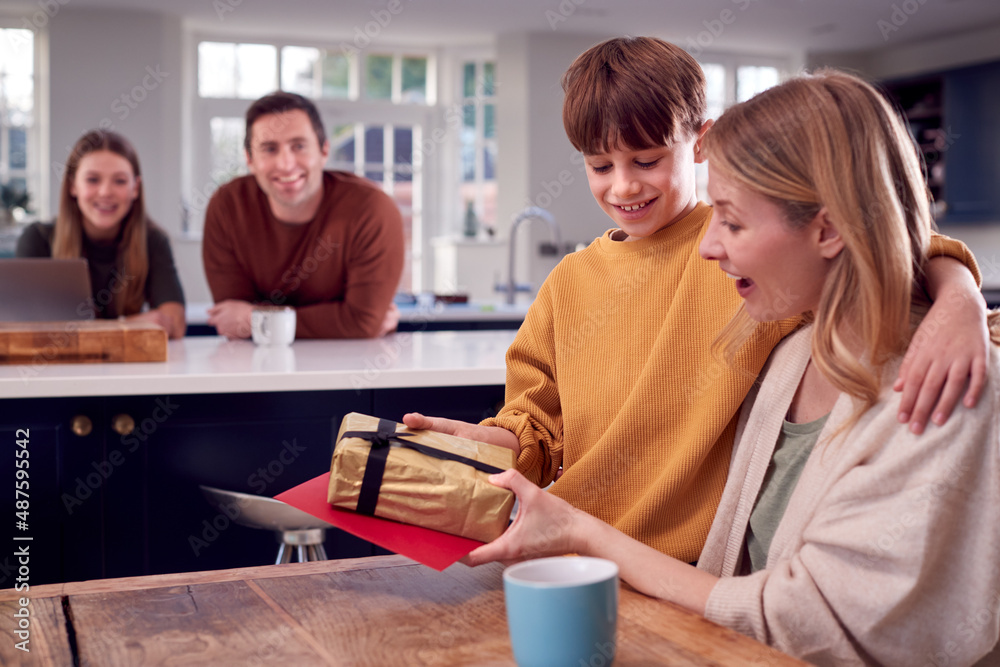 Son Giving Mother Gift As She Sits At Kitchen Table At Home Son Giving Mother Gift As She Sits At Kitchen Table At Home