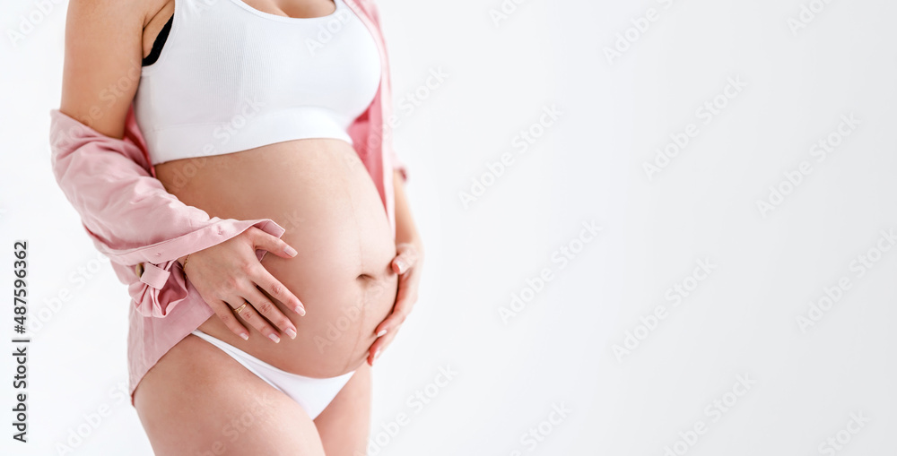 A pregnant woman in a pink shirt touches her big belly on a white background. The concept of motherhood, pregnancy and baby waiting. Copy space, banner
