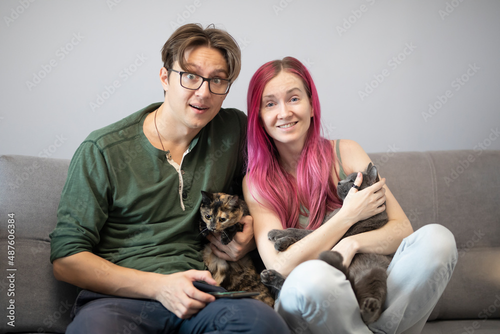 a man and a woman sit on a gray sofa and hug cats
