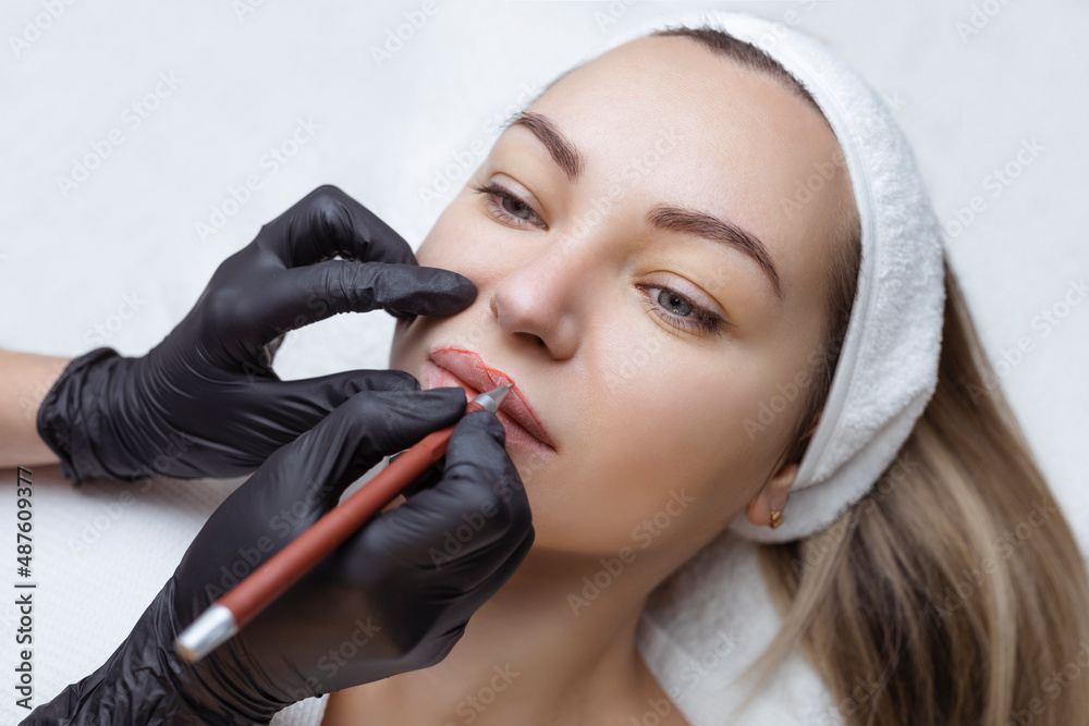 Young woman with permanent makeup on her lips in a cosmetologists salon. Permanent makeup tattoo. draw a line with a red lip pencil - obrazy, fototapety, plakaty 