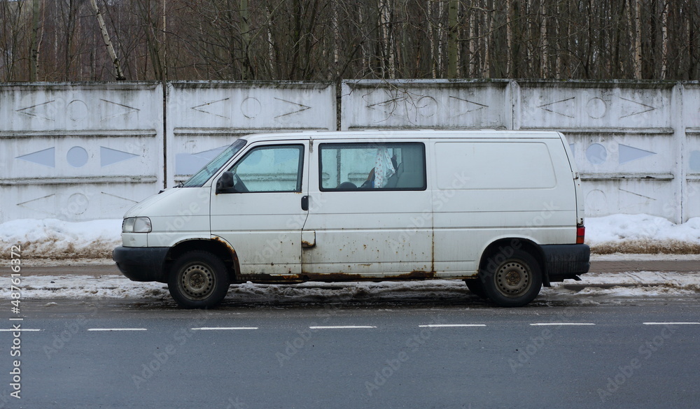 Old rusty white minivan parked at a gray concrete fence, Klochkov Lane, St. Petersburg, Russia, February 2022