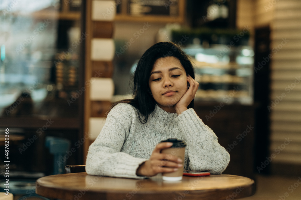 A dark-skinned young woman sits in a cafe and drinks coffee while waiting for her partner.