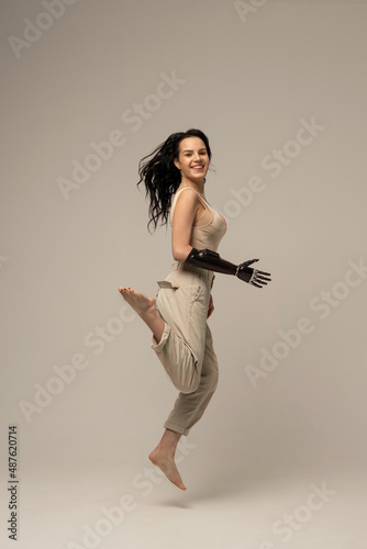 Woman with prosthetic robotic arm, bionic artificial hand jumping isolated on beige background. Natural Skin Nature Makeup Fresh Spa Women's Cosmetic Portrait. Beauty variety. Positive. Copy space