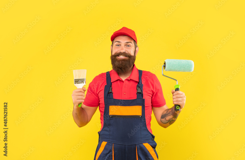 happy bearded man renovator in work clothes hold paint roller and brush on yellow background
