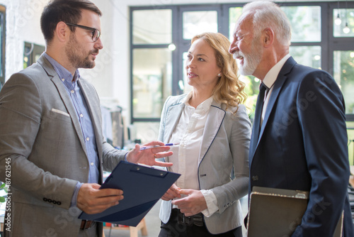 Group portrait of three happy positive business professionals at work. Team of confident people in smart suits standing in office and discussing document.