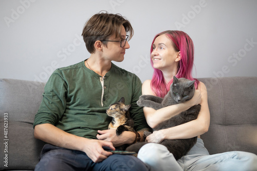 a man and a woman sit on a gray sofa and hug cats