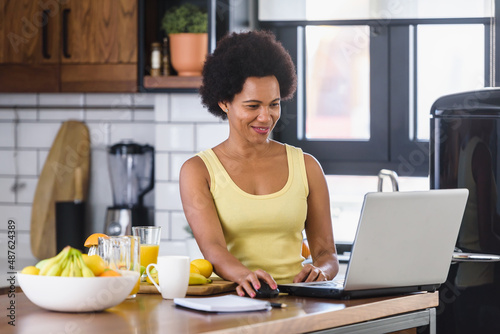 Young cheerful African-American woman surfing the internet on a laptop computer at home in the kitchen and refreshing with a fruit juice drink.
