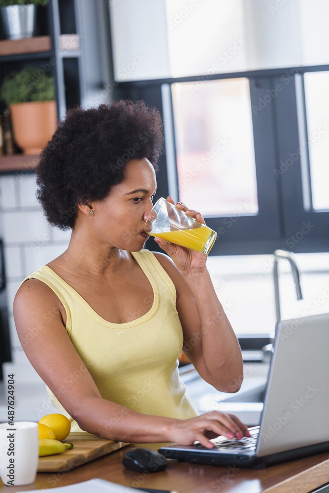 Young cheerful African-American woman surfing the internet on a laptop computer at home in the kitchen and refreshing with a fruit juice drink. Young cheerful African-American woman surfing the internet on a laptop computer at home in the kitchen and refreshing with a fruit juice drink.