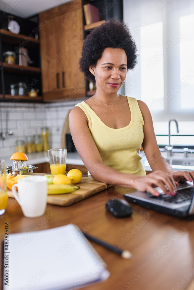 Young cheerful African-American woman surfing the internet on a laptop computer at home in the kitchen and refreshing with a fruit juice drink.