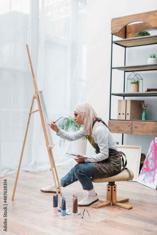 side view of muslim woman drawing on easel near paint tubes on floor.