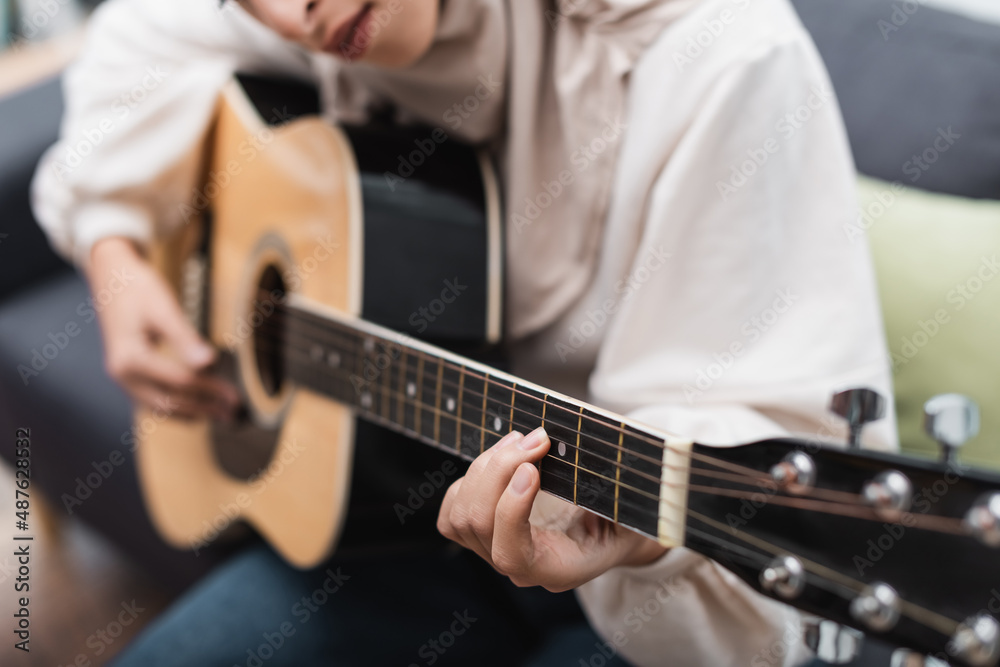 cropped view of blurred muslim woman playing acoustic guitar at home.