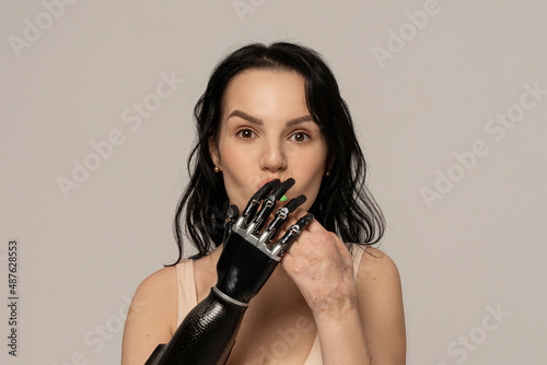 Beauty disabled Woman closing mouth by bionic arm, artificial hand isolated on beige background. Surprised. Natural Skin Makeup Fresh Spa Women's Cosmetic Portrait. Beauty variety. Copy space
