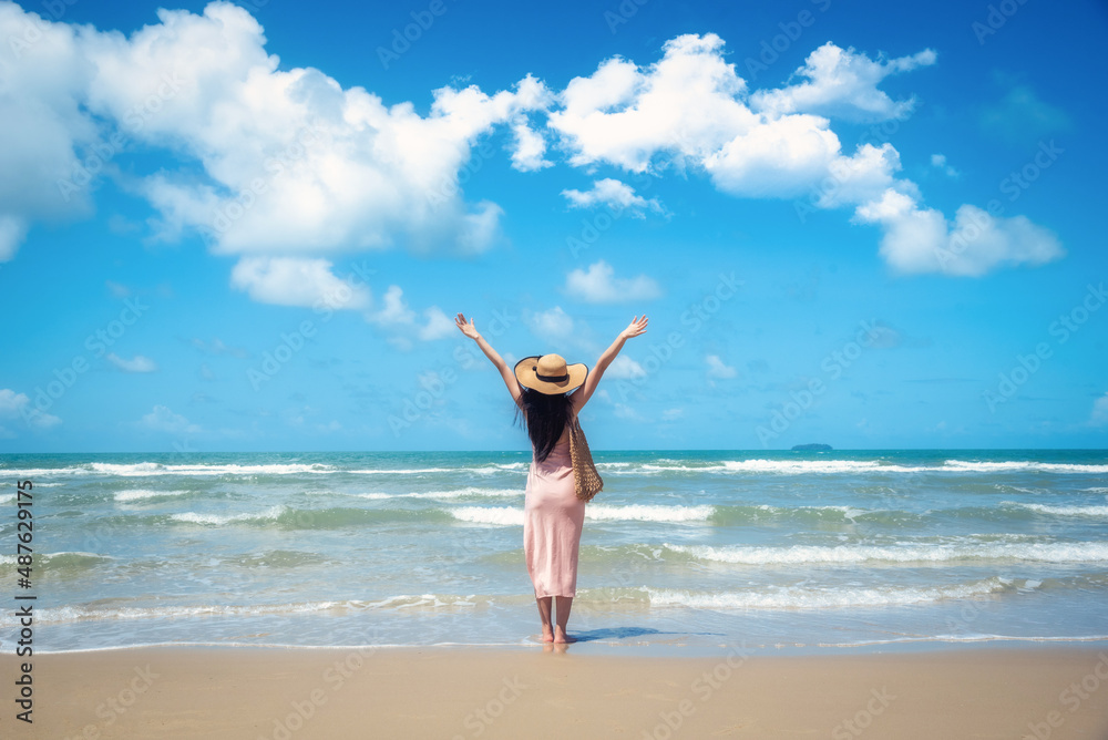 Happy traveler woman in sunhat and dress standing with her arms raised looking tropical beach on summer day. Holiday and summer travel concept Happy traveler woman in sunhat and dress standing with her arms raised looking tropical beach on summer day. Holiday and summer travel concept