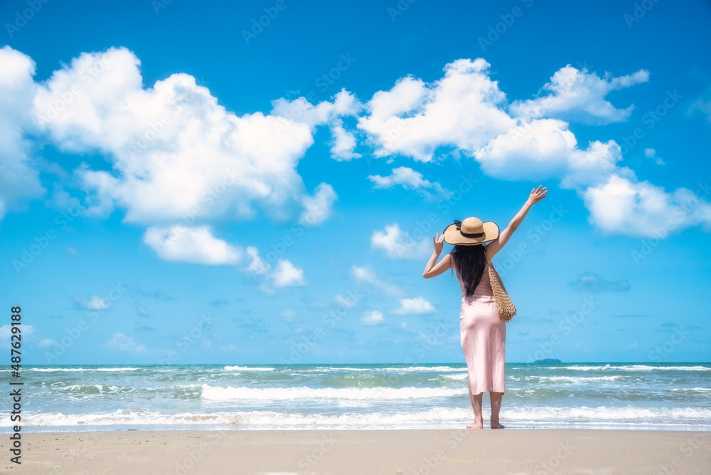 Happy traveler woman in sunhat and dress standing with her arms raised looking tropical beach on summer day. Holiday and summer travel concept Happy traveler woman in sunhat and dress standing with her arms raised looking tropical beach on summer day. Holiday and summer travel concept
