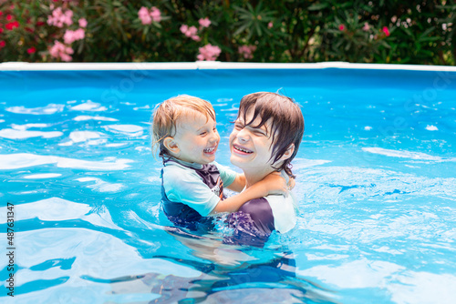 Two brothers of different age are playing in a swimming pool in summer during vacation. Happy siblings spending time together.