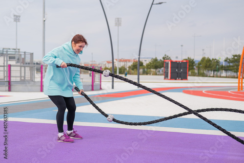 Caucasian woman in a mint sweatshirt is training with battle ropes at the sports ground. 