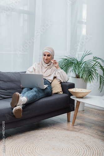 cheerful arabian woman in hijab eating popcorn and watching film on laptop on couch.