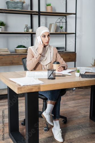 muslim woman talking on smartphone while sitting near coffee to go on work desk at home.