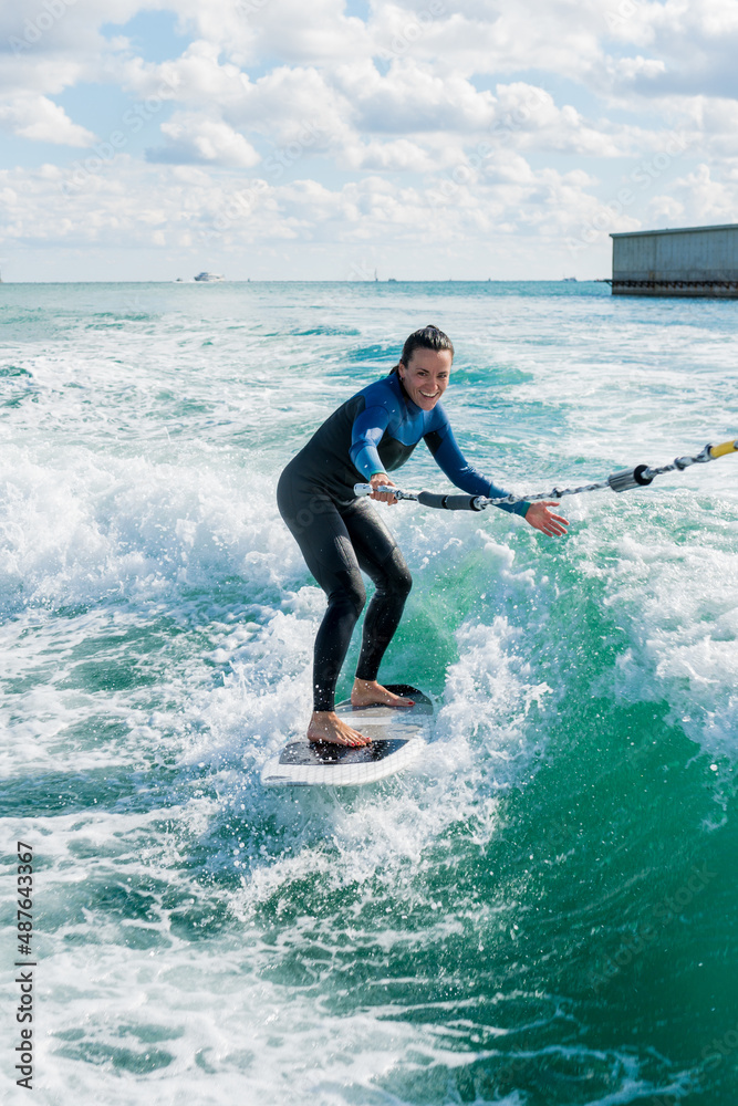 Female in wetsuit riding the waves using of tow rope behind a boat on sunny day. Young athletic woman learning wakesurfing and perfecting tricks. Watersport concept. Female in wetsuit riding the waves using of tow rope behind a boat on sunny day. Young athletic woman learning wakesurfing and perfecting tricks. Watersport concept.