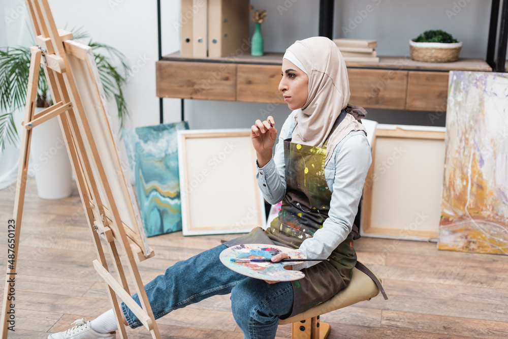 muslim woman with palette sitting near easel in home studio. muslim woman with palette sitting near easel in home studio.