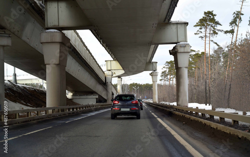 Cars are moving in a traffic jam under a trestle bridge in a forest belt with tall pines.
