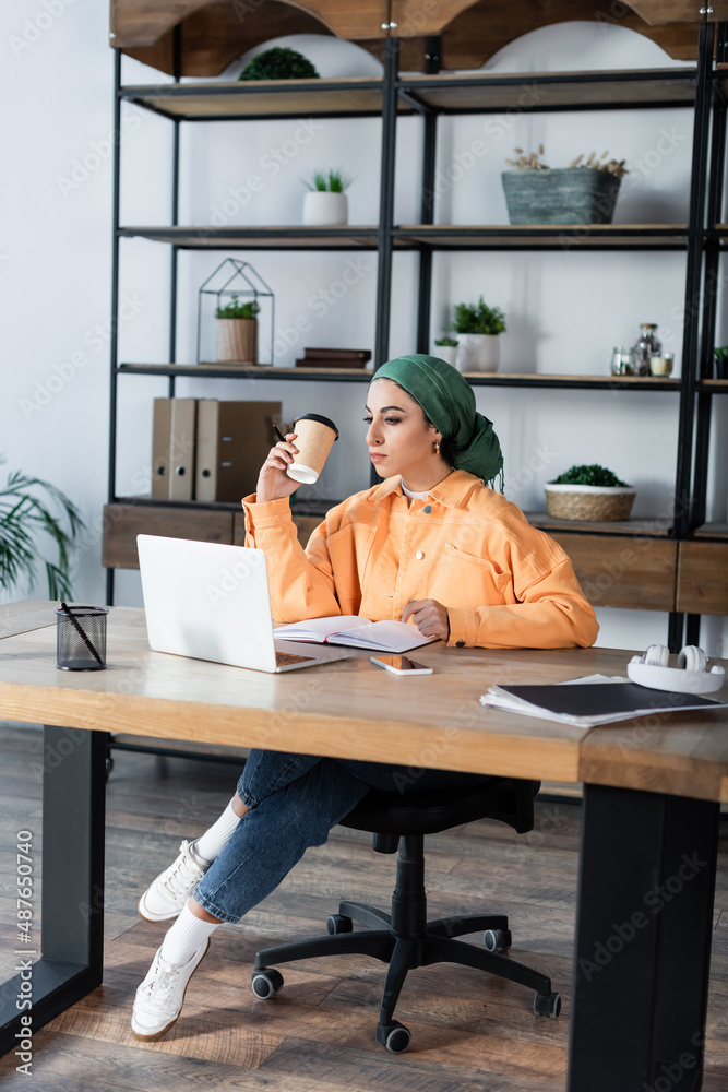 muslim woman holding paper cup while sitting near laptop at home.
