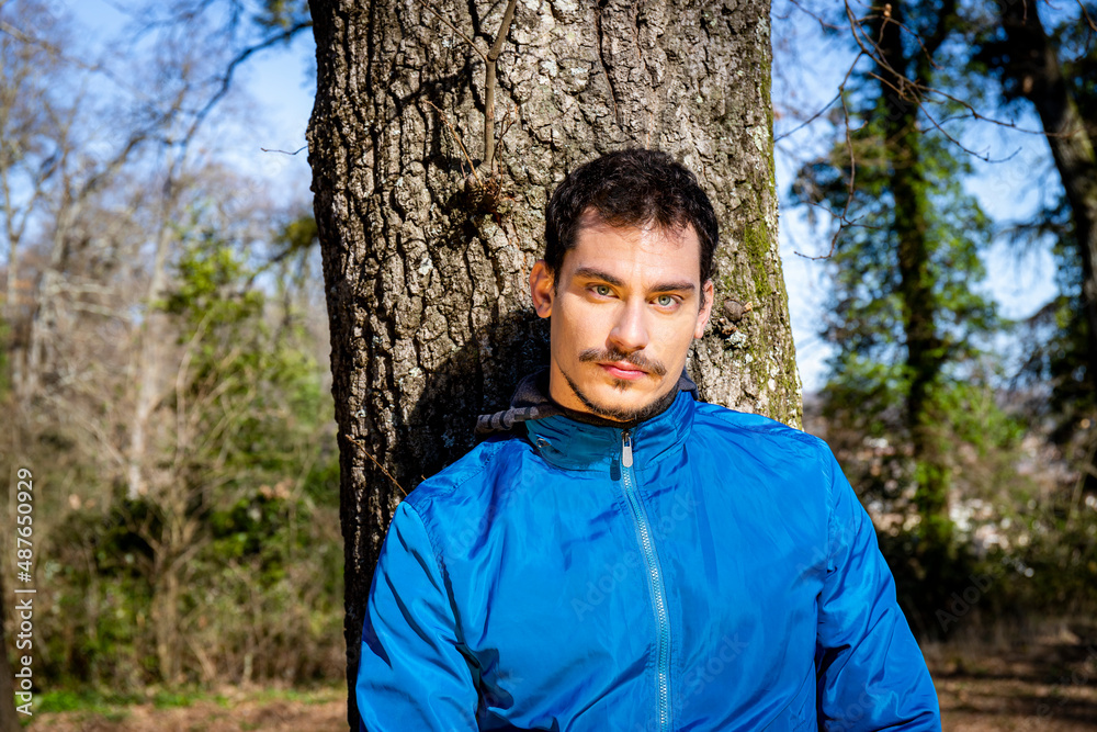 Portrait of a beautiful young man with blue eyes. The man is training in the woods and is leaning against an oak tree.