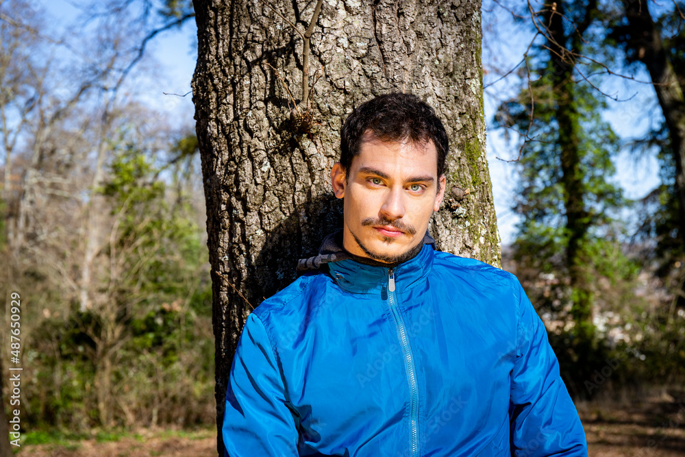 Portrait of a beautiful young man with blue eyes. The man is training in the woods and is leaning against an oak tree. Portrait of a beautiful young man with blue eyes. The man is training in the woods and is leaning against an oak tree.